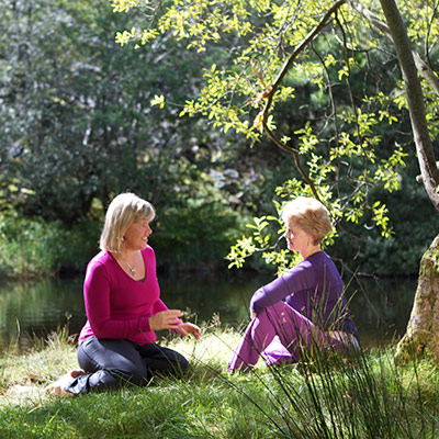 Two women chatting by a river under a tree