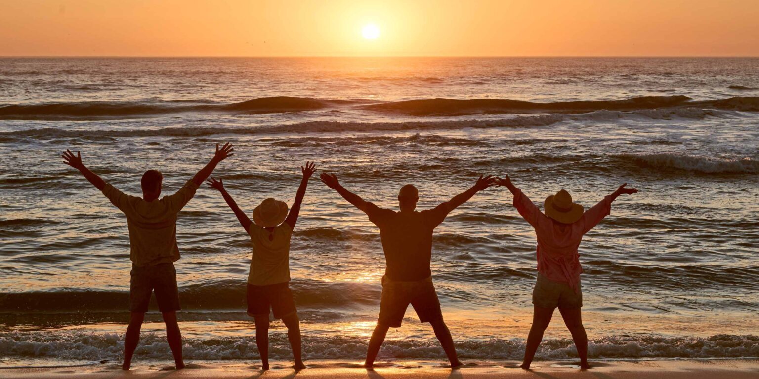 Silhouette Of group On Beach Looking Out To Sea At Sunset With Arms Outstretched